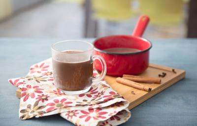 Fotografia em tons de vermelho e azul em uma bancada de madeira azul, uma tábua de madeira, um pano branco com flores rosas e marrons, uma panelinha vermelha pequena com o chocolate quente dentro dele. Ao centro, uma xícara de vidro com o chocolate quente